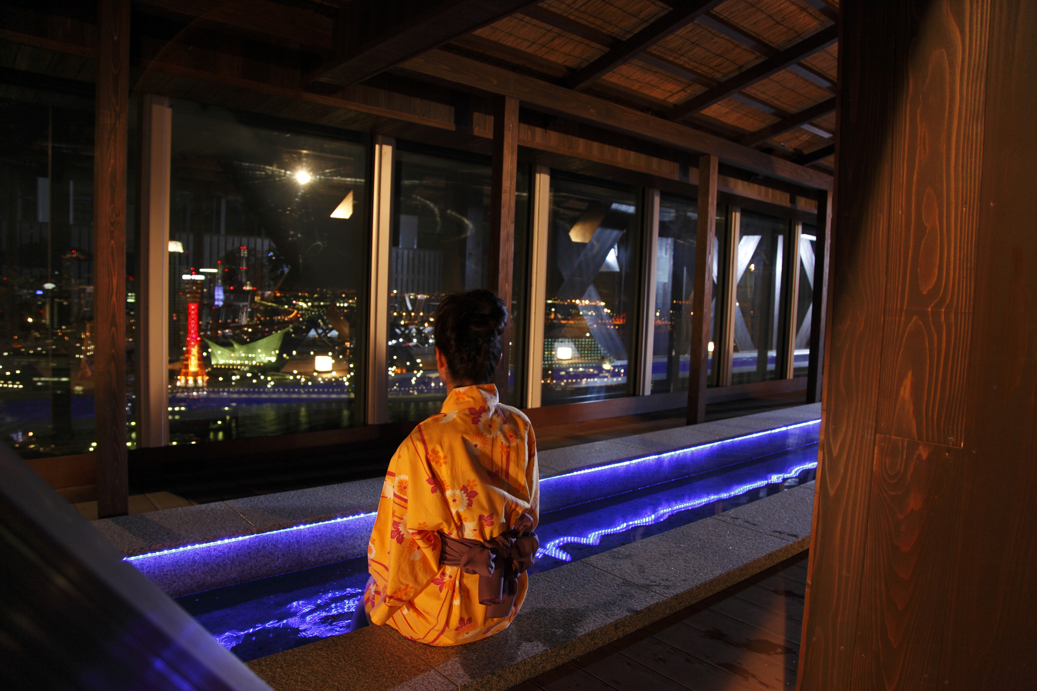 万葉倶楽部の屋上の足湯。窓の外に神戸港の夜景が広がり、浴衣の人が湯に足をつけている