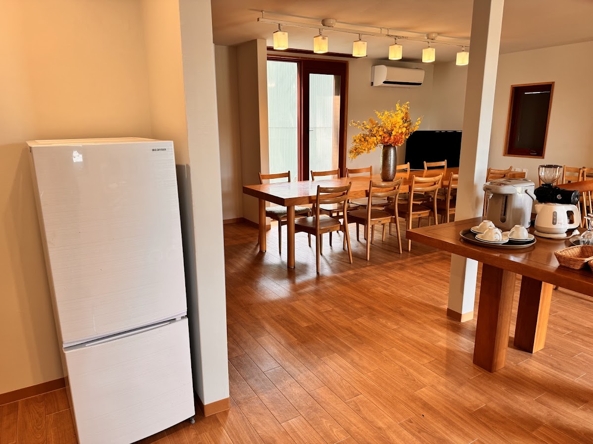 広々とした木のダイニングテーブルと、使いやすく整えられたキッチン設備が揃う薫風舎の共有スペース。炊飯器やトースター、コーヒーメーカーなどの家電に加え、食器類も完備されており、滞在者が自由に調理や食事を楽しめます。中央に飾られた季節の花が、空間に彩りと温もりを添え、旅のひとときを心地よく演出します。