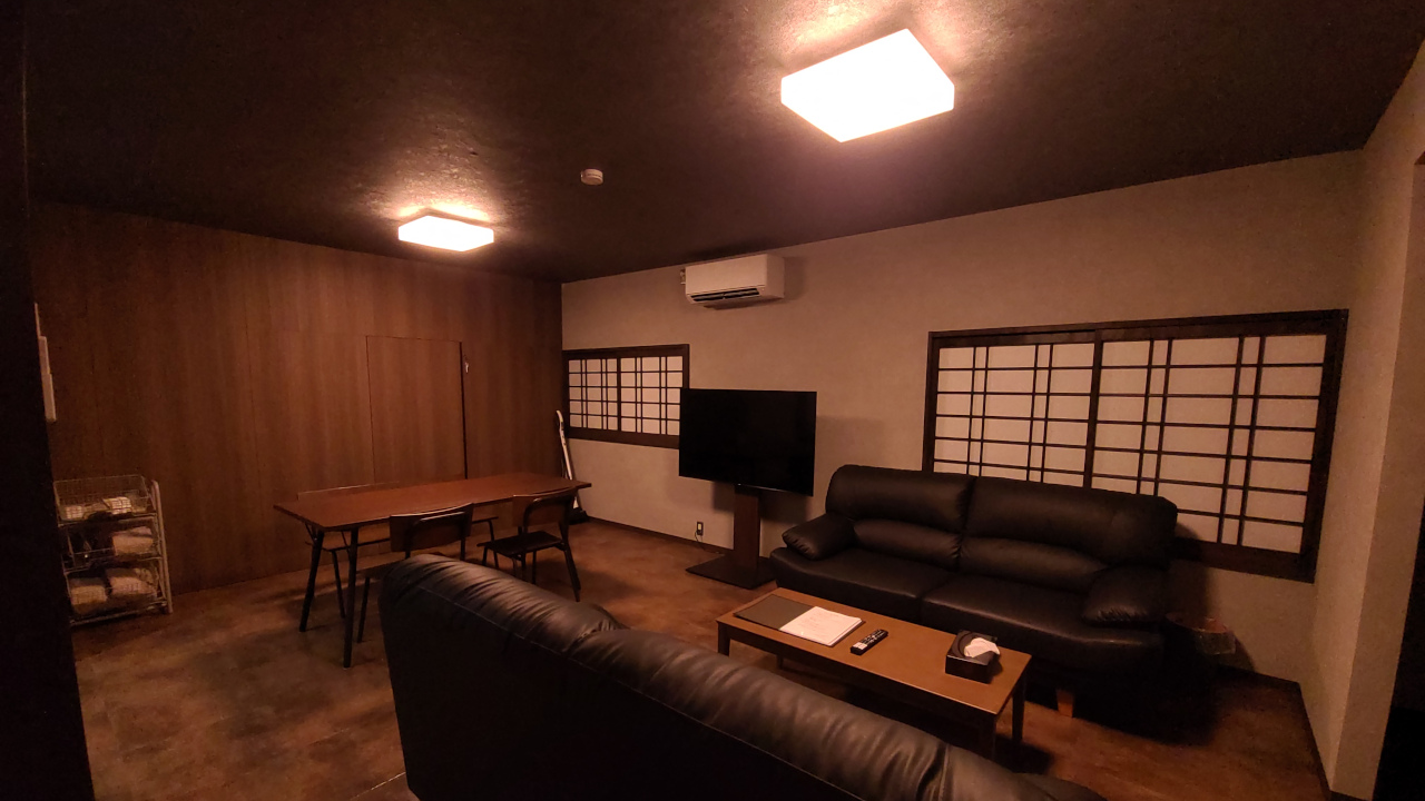 ダイニングテーブルとソファーの間の東側の壁際にテレビがあります。北西の壁際にはタオル等を設置したアメニティセットのかごを置いています。リビングの北側にはダイニングテーブルがあり、2人掛けのベンチシートと2つの1人掛けの椅子があり、全部で4人が座れます。リビングの南側には1つのローテーブルをはさんで2人掛けのソファーが2つあります。少し窮屈ですがソファー１つあたり1人が眠ることができます。